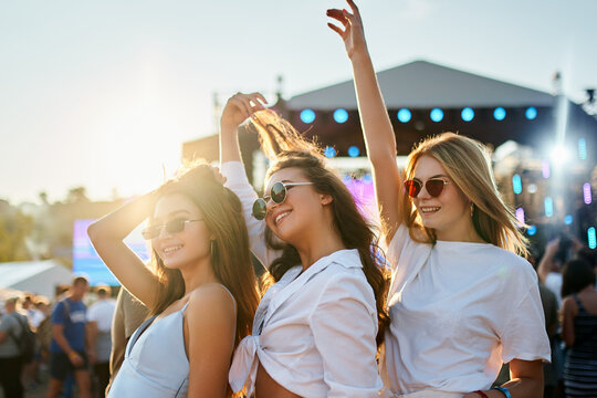 Girls in white tops enjoy summer music festival on sandy beach at sunset. Arms raised, they dance, laugh, celebrate friendship in bright sunlight, crowd, stage in background. Holiday, youth culture.