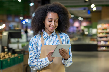 An employee with afro hair, wearing an apron, uses a tablet in a grocery store for inventory or customer service. This showcases efficient and friendly customer interaction in a modern market
