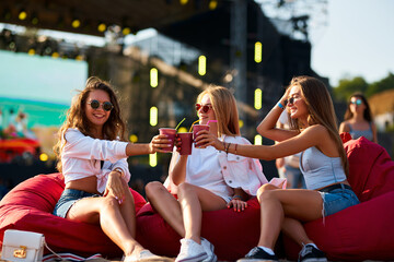 Friends clink drinks on red bean bag chairs at sunny beach fest. Girls in sunglasses enjoy music...