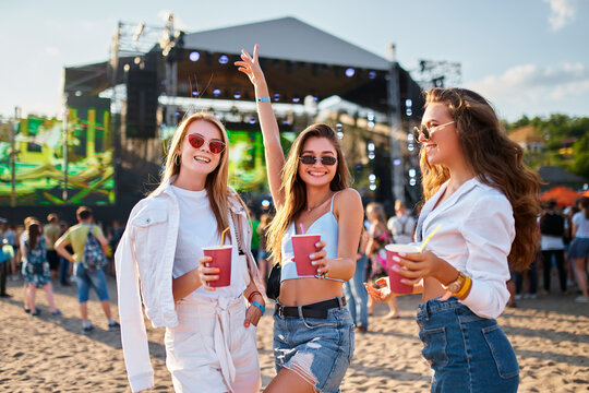 Girls toast with cold drinks at beach music fest in summer, smiling, wearing sunglasses, with stage, people in background. Celebration, fun, youth lifestyle. Females enjoying party, ocean nearby.