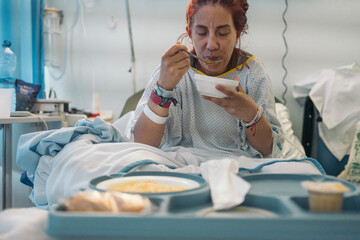 Redheaded woman on hospital bed blowing on spoonful of food from tray in a clinical setting.