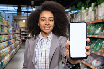 In a supermarket aisle, a young African American woman is displaying a smartphone with a blank...