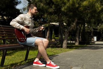 Attractive young guy playing guitar while sitting on a bench