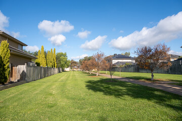 A public green grass lawn with a pedestrian walkway or footpath in a residential neighborhood with some houses. Background texture a vacant land lot in a suburb with two-story homes. 