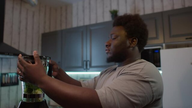 Overweight Black man closing lid of kitchen blender while going to make green smoothie drink for healthy dinner at home
