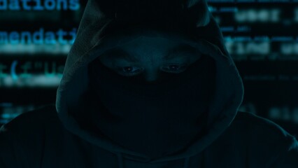 An anonymous hacker. Hooded figure with a masked face, program code in the background in a dark...