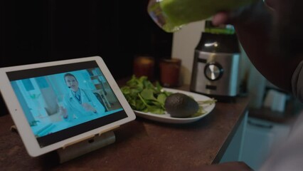 Close up shot of unrecognizable Black man drinking green smoothie and speaking with female dietician via video call on digital tablet while having online medical consultation