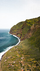 An exquisite drone capture showcases the verdant, vegetated cliffs of Madeira island, where they converge with the deep blue ocean waters on a sun-drenched day, with a charming village nestled nearby.