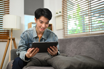 Smiling asian male freelancer sitting on couch and working with digital tablet