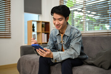 Satisfied young man holding credit card and using mobile phone making transaction shopping online