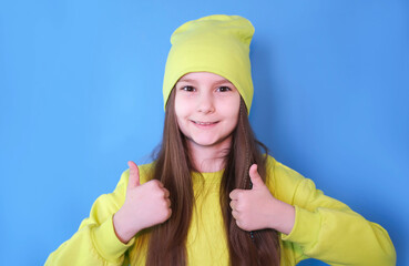 Little smiling girl showing thumbs up with hand on blue background with copy space.