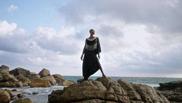 LGBTQ bipoc person in luxury dress, boutique golden jewelry poses against lighthouse on cliff, ocean, looks aside. Divine gender fluid multiethnic model in epic outfit stands on rock. Cloudy sky.