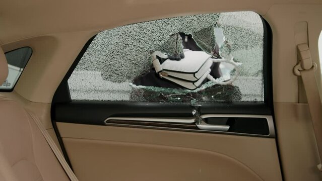 Thief's hands steal lady's handbag through broken side window vandalism act vehicle crime theft happening. Hands of thief reach through shattered side glass taking purse from car criminal activity.
