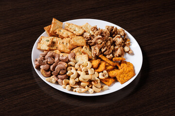 A plate with dry snacks - nuts and crackers on a dark table background. Angled view. Selective focus.