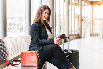 A female traveler looking at her phone, sitting in the waiting room of a train station, carrying...