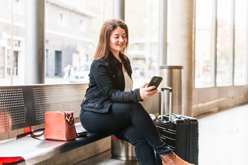 A female traveler smiles while looking at her phone, sitting in the waiting room of a train...