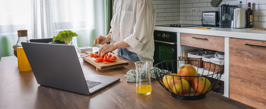 Woman standing in the kitchen preparing salad and chatting online using laptop