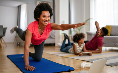 Young woman, mother exercising at home in living room, father playing with kid in background.