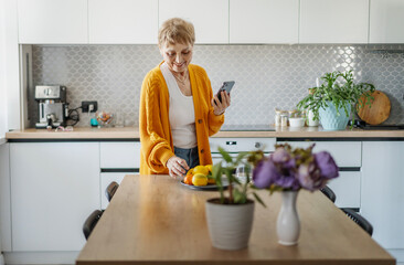 Senior happy Caucasian woman in a yellow cardigan standing in the kitchen with a smartphone in her...
