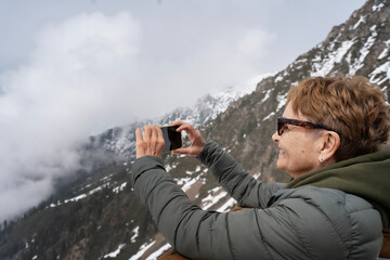 Attractive happy senior elderly woman travelling in the mountains taking a photo using a smartphone