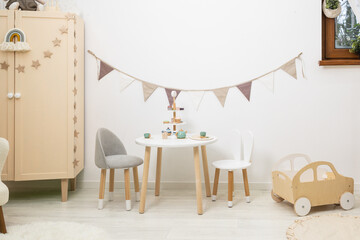 Scandinavian cozy interior children's room. Games table and chairs. Table wooden service waiting for tea and cakes. Wooden cabinet. Fabric garland flags. Wooden machine. Chair with ears