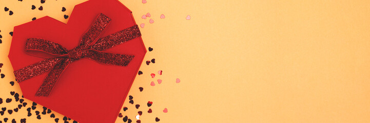 Banner with red gift box in a heart shape and confetti on a golden background.