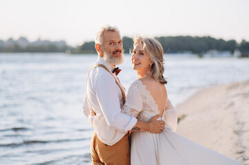 Senior newlyweds hug. Stylish couple of elderly newlyweds stand embracing on river bank.