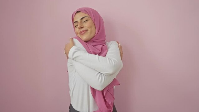 Beautiful young hispanic woman in hijab hugging self with joy over pink isolated background. portrait of happy muslim female smiling with confidence