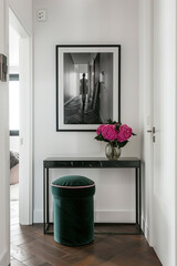 Fototapeta premium A small black console table with an emerald velvet stool is located in the center of an empty white wall. On it, there is a photo frame and pink peonies. The photo depicts a modern apartment interior