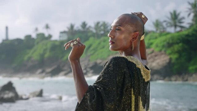Gorgeous gay black man in luxury gown poses on scenic ocean beach slowmo. Non-binary ethnic fashion model in long posh dress and accessories looks at camera, dances with hands in slow motion