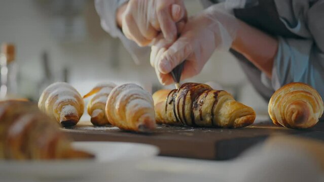 A talented pastry chef wearing protective gloves pours fresh chocolate over a croissant. Experienced worker pours dark chocolate on baked goods following sustainable business principles