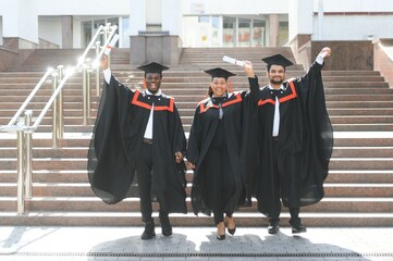 Group of happy smiling multicultural people in graduation gowns and caps with diplomas in hands...