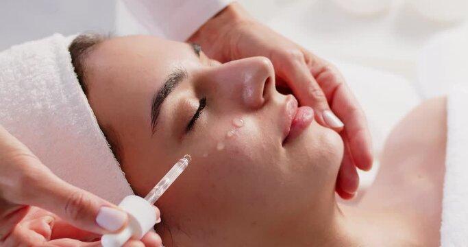 Woman spending time in spa salon, enjoying beauty treatments and relaxing. Skin care specialist uses dropper to apply cosmetic serum around eyes and on cheeks of pretty young lady with towel on head