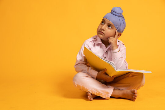 Indian sikh school kid sitting on floor holding note book thinking studying isolated over yellow background. student preparing for exams, Knowledge and intelligence, Punjab education