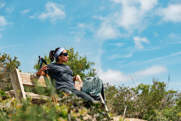 Hiking woman with glasses and mountain sticks, rests on a bench at the top.