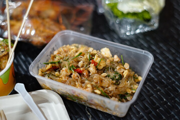 Stir-fried glass noodles in a clear plastic food container on a black rattan tab