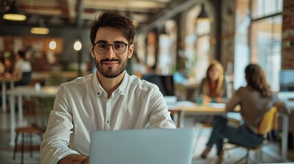 Young Man in Glasses and Shirt Working on Laptop in a Creative Business Agency. Diverse People in the Background. In Good Mood.