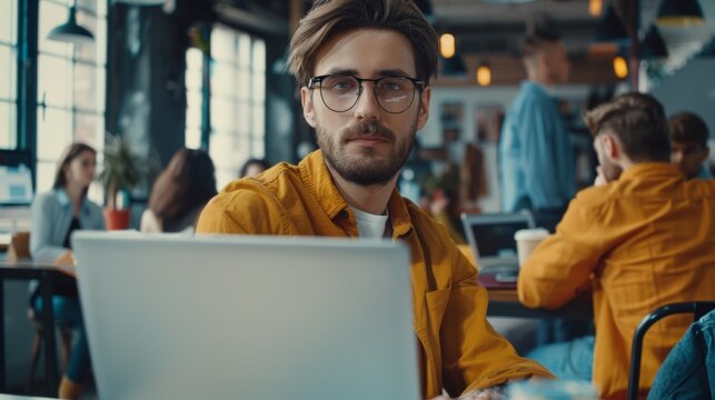 His mood is good. He is working on a laptop in a loft office of a creative business agency. People working in the background seem to be from different walks of life.