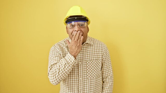 Shell-shocked middle-age latin man, hand covering mouth in surprise, security glasses and hardhat on, fear evident, stunned silent against isolated yellow background