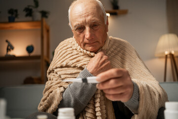 Close-up of elderly man wearing blanket and holding thermometer in one hand. Antipyretic. Viral...