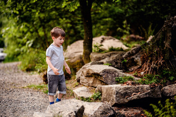 Young boy in a gray t-shirt and denim shorts exploring a gravel path near large rocks and trees....