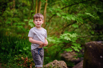A young boy smiles brightly while standing on a forest path surrounded by lush green foliage and rocks. He is enjoying a peaceful nature walk.