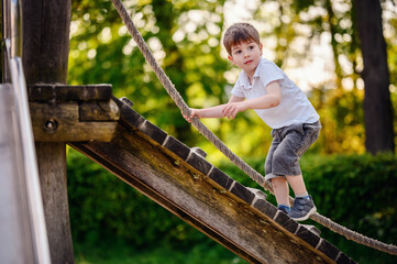 A young boy in a white shirt and denim shorts enthusiastically climbs a wooden ramp using a rope at...