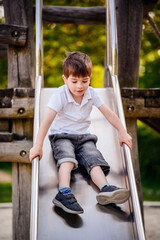A young boy in a white shirt and denim shorts slides down a metal playground slide with a concentrated expression, surrounded by green trees on a sunny day.