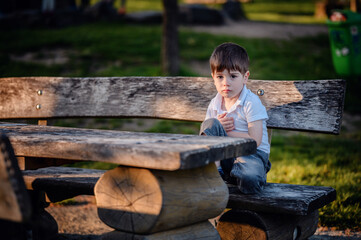 A young boy in a white shirt and denim shorts sits on a wooden bench in a park, looking thoughtful and surrounded by greenery on a sunny day.