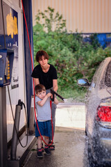 Mother helps her young son wash a car at a self-service station. They both hold the hose, spraying...