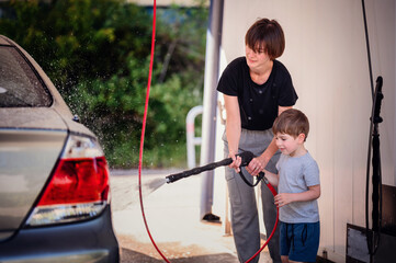 Smiling mother guiding her young son in using a high-pressure hose to wash a car at a self-service...
