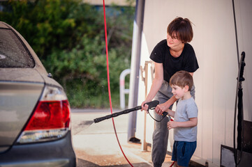 Smiling mother guiding her young son in using a high-pressure hose to wash a car at a self-service...