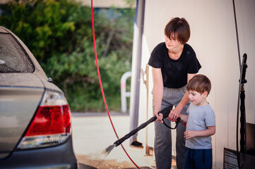 Smiling mother guiding her young son in using a high-pressure hose to wash a car at a self-service...