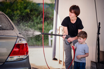 Smiling mother guiding her young son in using a high-pressure hose to wash a car at a self-service...